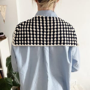 K&ouml;nnte beinhalten: Ein handgeh&auml;kelter Schal in einem Muster aus Marineblau und Creme, getragen &uuml;ber einem hellblauen Hemd. Der Schal hat einen cremefarbenen Rand. Das Bild zeigt die R&uuml;ckseite, die das Design und die Textur des Schals hervorhebt.
