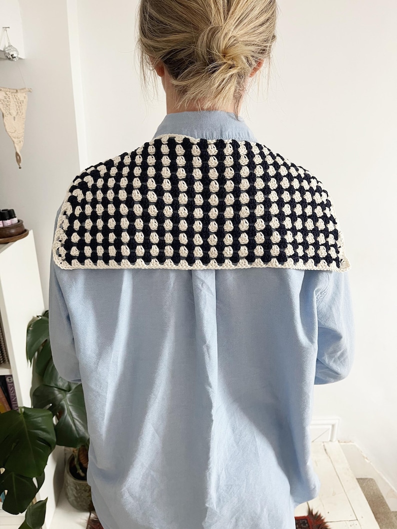 K&ouml;nnte beinhalten: Ein handgeh&auml;kelter Schal in einem Muster aus Marineblau und Creme, getragen &uuml;ber einem hellblauen Hemd. Der Schal hat einen cremefarbenen Rand. Das Bild zeigt die R&uuml;ckseite, die das Design und die Textur des Schals hervorhebt.