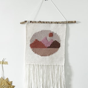 May include: A white crochet wall hanging with a landscape design. The design features a circular motif with brown, pink, and red mountains. The hanging is suspended from a wooden dowel and has white fringe.