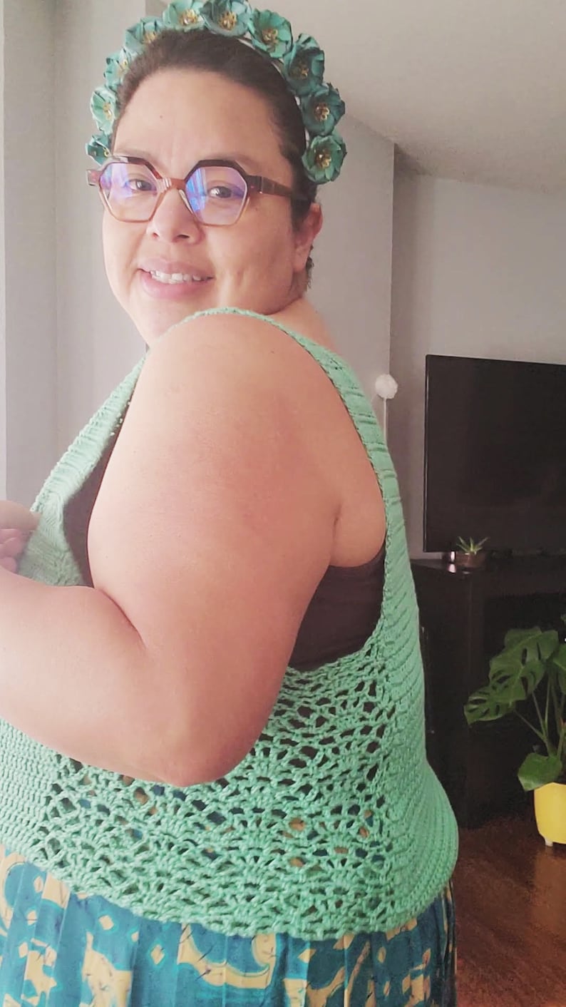 May include: A woman wearing a green crochet top with a floral headband. The top has a delicate openwork pattern and is worn over a patterned skirt.