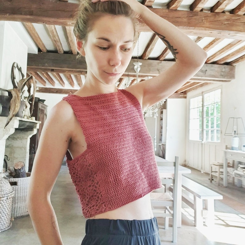May include: A close-up of a woman wearing a pink crocheted crop top with a square neckline and a delicate openwork pattern. The top is sleeveless and has a cropped length, showing the woman's midriff.