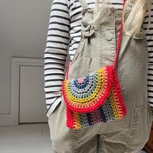 Könnte beinhalten: Ein Kind in Latzhose und gestreiftem Oberteil trägt eine Regenbogen-Garn-Umhängetasche. Die Tasche hat eine rot-orange-gelb-grün-blau-violette Farbgebung.