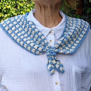K&ouml;nnte beinhalten: Ein blau-cremefarbener geh&auml;kelter Kragen, der vorne gebunden ist. Der Kragen besteht aus kleinen, quadratischen H&auml;kelstichen. Der Kragen wird &uuml;ber einem wei&szlig;en Hemd mit Kn&ouml;pfen getragen.