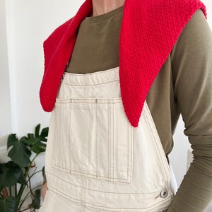 Peut inclure: Un ch&acirc;le en tricot rouge vif drap&eacute; sur les &eacute;paules. L'ensemble comprend une salopette en jean de couleur cr&egrave;me avec des coutures apparentes et un haut vert olive &agrave; manches longues. La salopette a des poches. L'ensemble est d&eacute;contract&eacute; et &eacute;l&eacute;gant.