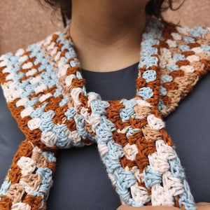 K&ouml;nnte beinhalten: Handgefertigter H&auml;kelschal im Granny-Square-Muster. Der Schal hat eine Farbpalette aus Hellblau, Braun, Beige und Wei&szlig;. Der Schal ist um den Hals gewickelt und aus weichem Garn gefertigt.