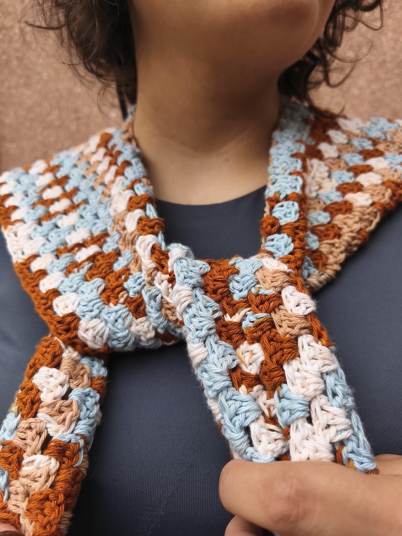 K&ouml;nnte beinhalten: Handgefertigter H&auml;kelschal im Granny-Square-Muster. Der Schal hat eine Farbpalette aus Hellblau, Braun, Beige und Wei&szlig;. Der Schal ist um den Hals gewickelt und aus weichem Garn gefertigt.
