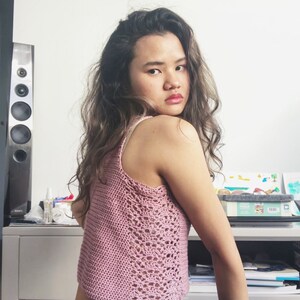 May include: A woman wearing a pink crocheted tank top and blue jeans. She is sitting on a chair with her back to the camera and looking over her shoulder.