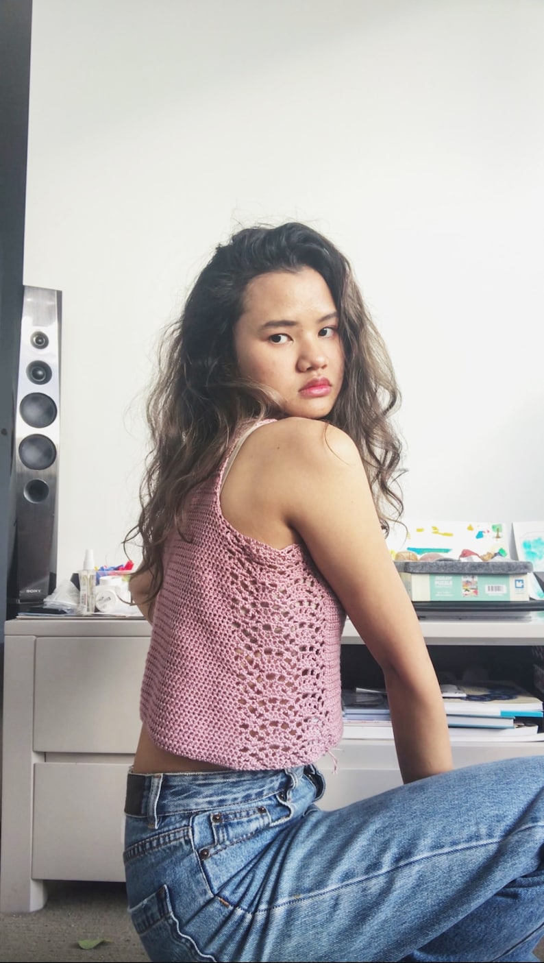 May include: A woman wearing a pink crocheted tank top and blue jeans. She is sitting on a chair with her back to the camera and looking over her shoulder.