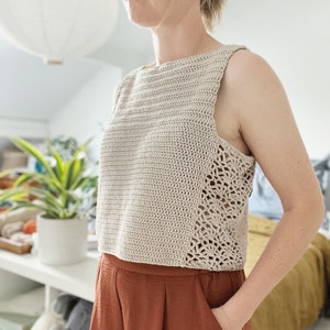 May include: A beige crochet top with a sleeveless design and a crocheted lace trim. The top has a relaxed fit and a cropped length.