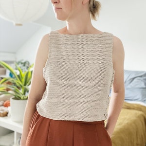 May include: A beige crochet tank top with a square neckline and a loose fit. The top is worn with rust-colored pants.