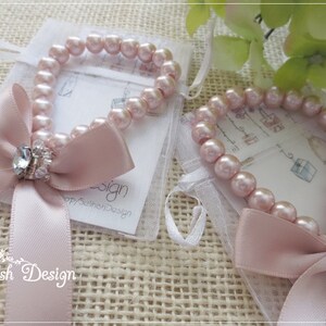 May include: Two pink pearl bracelets with a satin bow and a crystal accent. Each bracelet is presented in a small white mesh bag. The bracelets are on a textured, beige surface. The brand name "Selinish Design" is visible.