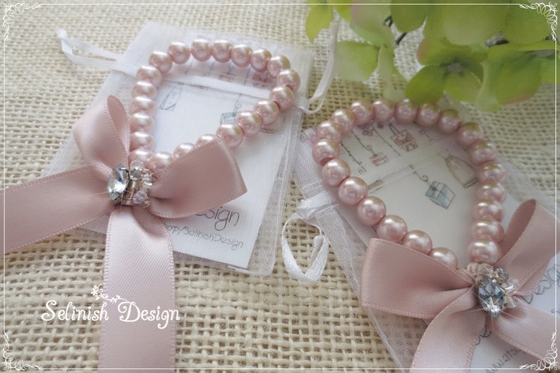 May include: Two pink pearl bracelets with a satin bow and a crystal accent. Each bracelet is presented in a small white mesh bag. The bracelets are on a textured, beige surface. The brand name "Selinish Design" is visible.