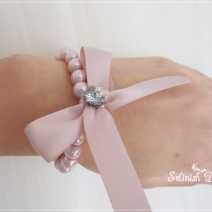 May include: A light pink pearl bracelet with a matching satin ribbon bow and a sparkling crystal centerpiece. The bracelet is designed to be worn on the wrist. The text "Selinish Design" is visible in the lower right corner.