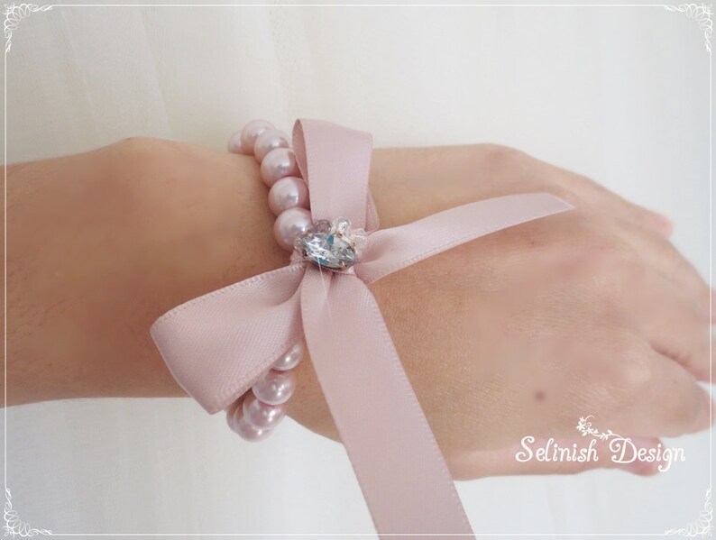 May include: A light pink pearl bracelet with a matching satin ribbon bow and a sparkling crystal centerpiece. The bracelet is designed to be worn on the wrist. The text "Selinish Design" is visible in the lower right corner.
