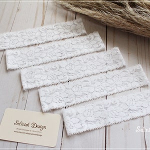 May include: Five white lace trim pieces with a floral pattern. The trim is approximately 1.5 inches wide. The trim is on a light wood surface. The text "Selinish Design" is on a business card in the lower right corner of the image.