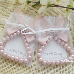 May include: Two pink pearl bracelets are displayed in sheer white pouches, each tied with a pink ribbon bow. The bracelets are arranged on a textured, light brown surface, with floral accents in the background. The text "Selinish Design" is visible.