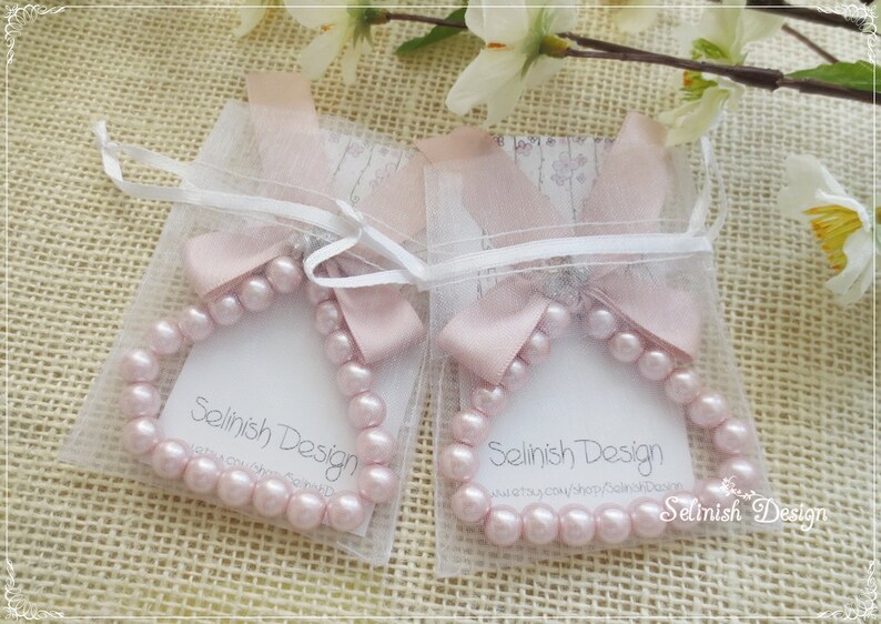May include: Two pink pearl bracelets are displayed in sheer white pouches, each tied with a pink ribbon bow. The bracelets are arranged on a textured, light brown surface, with floral accents in the background. The text "Selinish Design" is visible.