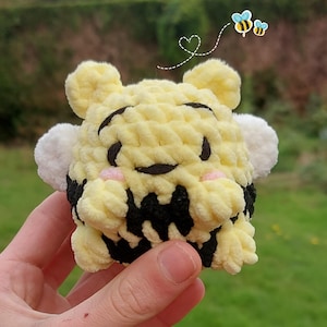 Op de afbeelding: Een klein, rond, gehaakt knuffeltje in de vorm van een bij met gele en zwarte strepen, witte vleugels en roze wangen. Het knuffeltje heeft geborduurde zwarte ogen en een mond, met een kleine afbeelding van twee bijtjes en een hartje erboven.