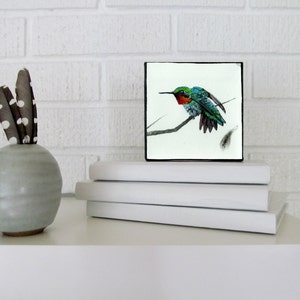May include: A framed watercolor painting of a hummingbird perched on a branch. The hummingbird is green, blue, and red with a long beak. The background is white.