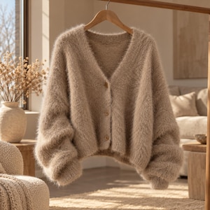 May include: A beige V-neck cardigan made of a fuzzy, soft-textured knit material. The garment features long sleeves with dropped shoulders and a front button closure with three matching buttons. The hem and cuffs are finished with a ribbed knit edge.