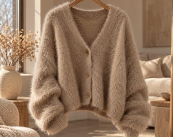 Peut inclure: Un cardigan beige à col en V confectionné dans une maille duveteuse et douce. Le vêtement présente des manches longues aux épaules tombantes et une fermeture boutonnée sur le devant avec trois boutons assortis. Les bords sont côtelés.