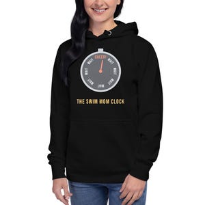 Puede incluir: Una sudadera con capucha negra que presenta un gráfico de una esfera de reloj circular con la palabra WAIT repetida alrededor del perímetro y la palabra CHEER! en la parte superior. Debajo, el texto THE SWIM MOM CLOCK está impreso en amarillo.