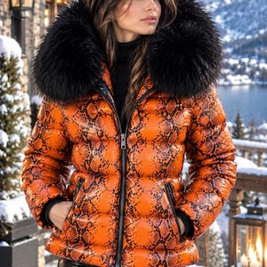 May include: A bright orange puffer jacket featuring a black snakeskin print pattern, a full-length front zipper, and a large, plush black faux fur hood trim. The jacket includes two side pockets with black zippers and black ribbed cuffs.