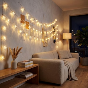 May include: A string of warm white star-shaped LED lights draped across a wall, illuminating a living room space. The lights feature multiple small, translucent stars connected by a thin wire, casting a soft glow over a sofa and decor.