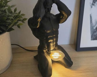 Könnte beinhalten: Eine mattschwarze Tischleuchte in Form einer knienden, muskulösen Figur mit den Händen hinter dem Kopf. Die Leuchte verfügt über eine warm leuchtende Edison-Glühbirne im Schrittbereich, die mit einem schwarzen Stromkabel verbunden ist.
