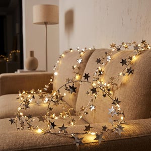 May include: A string of silver star-shaped ornaments intertwined with warm white LED fairy lights. The metallic stars are attached to a thin, flexible wire, creating a glowing, decorative garland that rests across the textured fabric surface.