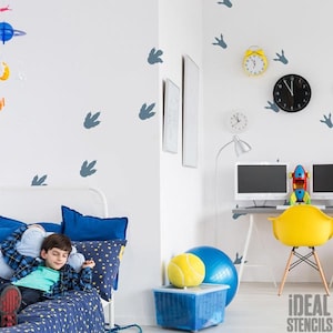 May include: A child's bedroom with dinosaur footprint wall decals. The room features a white bed with blue bedding, a yellow chair, and a desk with computer monitors. A yellow tennis ball and blue exercise ball sit on a blue storage bin.