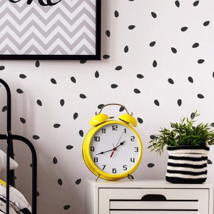 May include: A yellow alarm clock with black numbers and hands on a white nightstand with a black drawer. The clock is in front of a white wall with black seed-shaped decals. A green plant in a black and white striped pot is on the nightstand.
