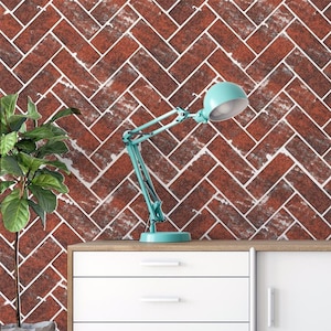Może przedstawiać: Turkusowa lampa biurkowa z regulowanym ramieniem stoi na drewnianej powierzchni nad białą szafką z trzema szufladami i drzwiami. Tło to ściana z wzorem cegieł w odcieniach czerwieni i bieli. Po lewej stronie znajduje się roślina doniczkowa.