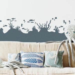 May include: A white couch with a gray and white octopus plush toy and pillows with a blue and white fish pattern. The wall behind the couch has a gray silhouette of an underwater scene with fish, coral, and bubbles.