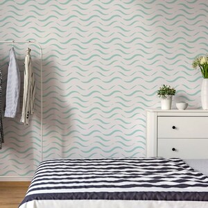 Può includere: Una camera da letto con una parete bianca con una carta da parati a motivi ondulati blu chiaro. Una cassettiera bianca con due cassetti si trova sul lato destro della stanza. Un letto con un piumone a strisce bianche e nere si trova in primo piano. Un appendiabiti bianco con diversi indumenti appesi si trova sul lato sinistro della stanza.
