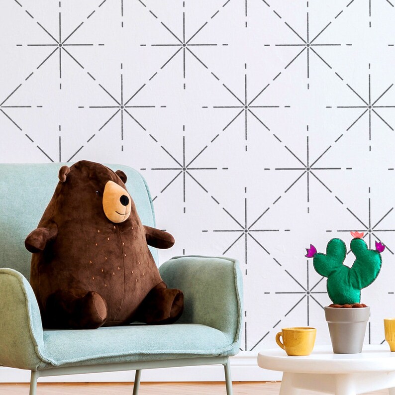 May include: A brown plush bear sits on a light blue chair. A green felt cactus with pink flowers is in a gray pot. A yellow mug sits on a white table. The wall has a geometric pattern.