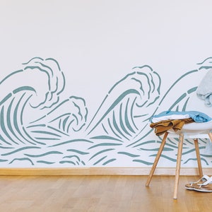 May include: A white chair with wooden legs sits in front of a white wall with a blue wave stencil design. The chair has a blue and brown blanket draped over it.