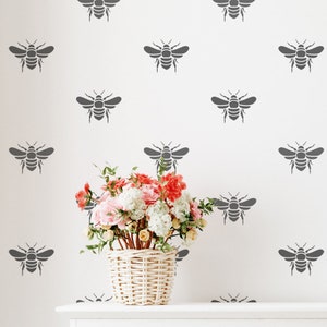 May include: A white wall with a repeating pattern of gray bee silhouettes. A wicker basket filled with pink and white flowers sits on a white shelf in front of the wall.