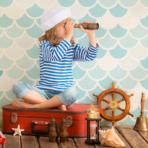 May include: A young person dressed as a sailor sits on a red suitcase with a nautical theme. They are holding a telescope and looking out at the horizon. The background is a blue and white fish scale pattern.
