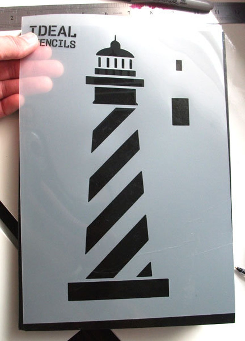 May include: A black and white lighthouse stencil with a striped design. The stencil is on a gray background and has the text "IDEAL STENCILS" in the top left corner.
