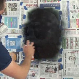 May include: A person wearing a respirator mask and a blue shirt is spray painting a black surface. The surface is covered with newspaper and blue painter's tape.