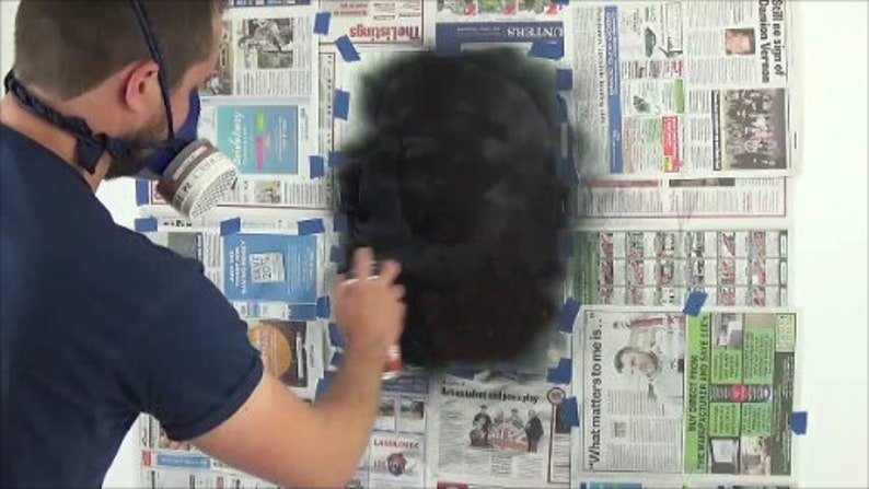 May include: A person wearing a respirator mask and a blue shirt is spray painting a black surface. The surface is covered with newspaper and blue painter's tape.