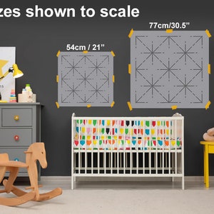May include: Two gray stencils with a geometric starburst design, taped to a dark gray wall. The stencils are labeled with sizes: 21 inches and 30.5 inches. The text "sizes shown to scale" is at the top. A crib and children's furniture are in the foreground.