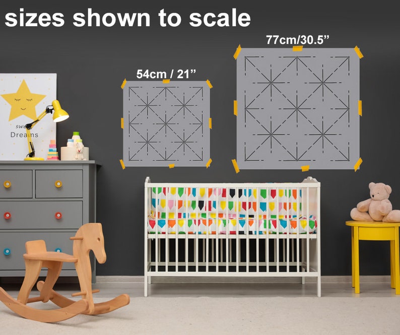 May include: Two gray stencils with a geometric starburst design, taped to a dark gray wall. The stencils are labeled with sizes: 21 inches and 30.5 inches. The text "sizes shown to scale" is at the top. A crib and children's furniture are in the foreground.