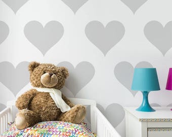 May include: A white wall with a gray heart pattern. A brown teddy bear sits on a white crib with a colorful patterned sheet. A blue lamp sits on a white nightstand.