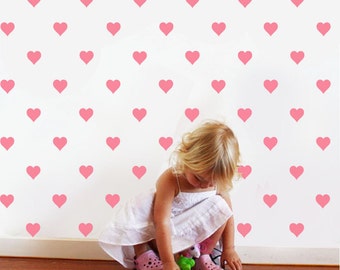 May include: A young child in a white dress is playing in front of a wall covered in pink heart stickers.