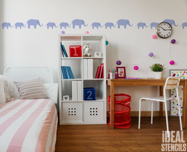 May include: A white bed with pink and white striped bedding, a white bookshelf with books and toys, a wooden desk with a chair, and a wall with a border of blue elephant stencils.