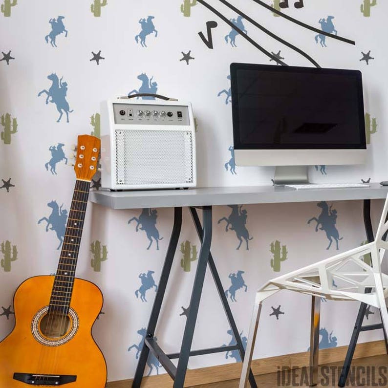 May include: A white guitar amplifier with a black knob and a black and brown acoustic guitar on a white desk with a black and gray chair and a computer monitor. The wall behind the desk is white with a blue and green pattern of cowboys riding horses and cacti.