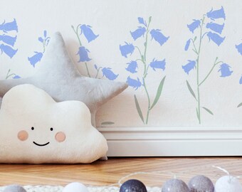 Puede incluir: Una pared blanca con un diseño de esténcil de flores azules. El esténcil presenta una fila de flores azules en forma de campana con tallos verdes. En primer plano, hay dos almohadas de felpa, una con forma de nube y la otra con forma de estrella.