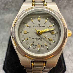 Può includere: Un orologio da polso bicolore argento e oro con quadrante argentato e lancette nere. Il quadrante dell'orologio ha un anello bianco attorno al bordo con numeri e segni neri. Il quadrante dell'orologio ha anche un anello bianco più piccolo con numeri e segni neri. Il quadrante dell'orologio ha il testo "DJ DAVID JORDAN" e "WATER 50MRESIST QUARTZ". L'orologio ha un cinturino in metallo argentato con accenti dorati.